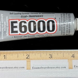 May include: A silver tube of E6000 industrial strength adhesive, labeled "Self-Leveling Formula / Fórmula de autonivelación" and "CLEAR - TRANSPARENTE". The tube is 1.0 fl oz (29.5 mL) and has a white cap. The tube is lying on a wooden ruler.