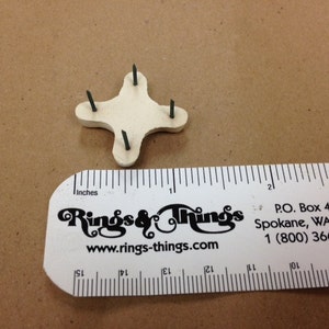 May include: A cream-colored, four-pointed object with four dark green pins, next to a ruler marked in inches. The ruler displays the text "Rings & Things" and a website address.