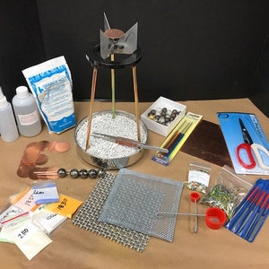 Puede incluir: Una colección de suministros para la fabricación de joyas, que incluye un quemador de trípode, discos de cobre, bolsas de polvo, botellas, malla, cuentas, limas, tijeras y otras herramientas. Los artículos están dispuestos sobre una superficie marrón.