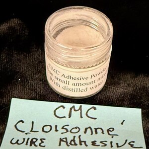 Op de afbeelding: Een klein, helder glazen potje gevuld met een fijn, gebroken wit poeder. Het potje heeft een wit etiket met zwarte tekst met de tekst "CMC Adhesive Powder" en instructies. Onder het potje staat een blauwe notitie met de tekst "CMC Cloisonne Wire Adhesive".