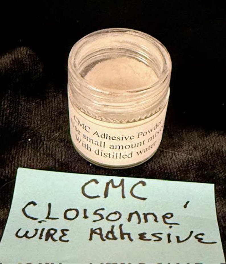 Op de afbeelding: Een klein, helder glazen potje gevuld met een fijn, gebroken wit poeder. Het potje heeft een wit etiket met zwarte tekst met de tekst "CMC Adhesive Powder" en instructies. Onder het potje staat een blauwe notitie met de tekst "CMC Cloisonne Wire Adhesive".