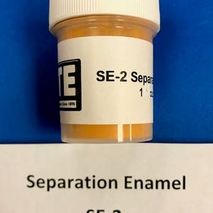 May include: A small, cylindrical jar with a white lid and a label displaying the text "SE-2 Separation Enamel." The jar contains a light orange powder. The label also includes the text "1 oz Dry."
