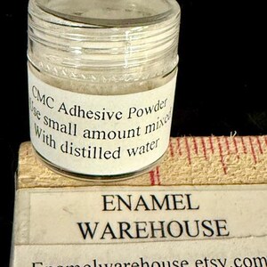 Op de afbeelding: Een klein, helder glazen potje gevuld met wit poeder, met het label "CMC Adhesive Powder". Het label instrueert om een kleine hoeveelheid te mengen met gedestilleerd water. Onder de pot staat een label met "ENAMEL WAREHOUSE" en de Etsy shop URL.