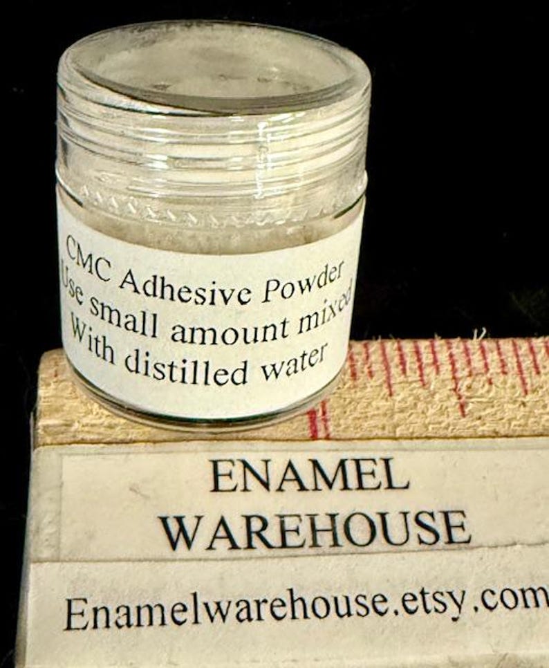 Op de afbeelding: Een klein, helder glazen potje gevuld met wit poeder, met het label "CMC Adhesive Powder". Het label instrueert om een kleine hoeveelheid te mengen met gedestilleerd water. Onder de pot staat een label met "ENAMEL WAREHOUSE" en de Etsy shop URL.