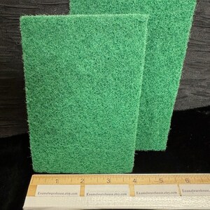 May include: Two green scrubby sponges, each measuring approximately 4 inches in length, are stacked on top of each other. The sponges are made of a soft, textured material and are ideal for cleaning dishes or other surfaces.
