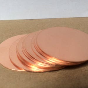 May include: A stack of copper-colored, circular metal discs. The discs are arranged in a slightly fanned-out pattern, revealing their edges. The surfaces of the discs reflect light, creating highlights. The background is a neutral tan color.