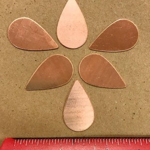 Puede incluir: Seis piezas de metal de cobre en forma de lágrima dispuestas en forma de flor sobre una superficie marrón.