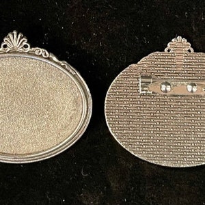 May include: A silver oval-shaped brooch with a decorative border. The back of the brooch has a pin and a textured surface.