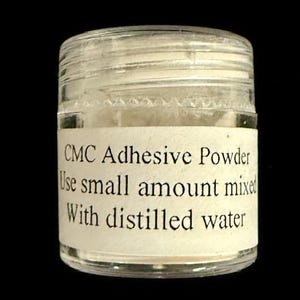 Op de afbeelding: Een kleine, heldere glazen pot gevuld met wit poeder. De pot heeft een wit etiket met zwarte tekst: "CMC Adhesive Powder. Use small amount mixed With distilled water."
