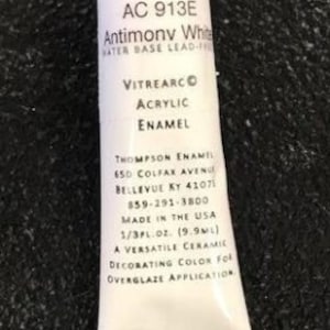 May include: A tube of Antimony White Vitrearc Acrylic Enamel paint. The tube is white with black text that reads "AC 913E", "Antimony White", "VITREARC Acrylic Enamel", "Thompson Enamel", "650 Colfax Avenue", "Bellevue KY 41071", "859-291-3800", "Made in the USA", "1/3FL.OZ. (9.9ML)", "A Versatile Ceramic Decorating Color for Overglaze Application".
