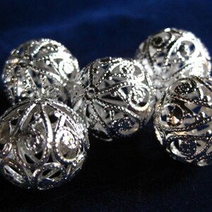 Puede incluir: Cinco cuentas de metal plateado con un diseño de filigrana decorativo. Las cuentas son redondas y tienen un centro hueco.