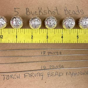 May include: Five silver buckshot beads, each about 1 inch in diameter, are lined up on a yellow ruler. The ruler is marked with inches and numbered 1 through 5. Below the ruler are two silver metal rods labeled "18 gauge" and "16 gauge". The text "TORCH FIRING BEAD MANDRELS" is handwritten below the rods.