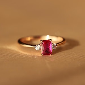 Op de afbeelding: Een goudkleurige ring met een rechthoekige, helderrode edelsteen als middelpunt. Twee kleinere, heldere edelstenen flankeren de hoofdsteen. De ring is geplaatst tegen een warme, neutrale achtergrond, wat de elegantie van het sieraad benadrukt.