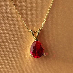 May include: A gold necklace featuring a teardrop-shaped, faceted red gemstone pendant. The pendant hangs from a delicate gold chain. The background is a solid, warm, golden-brown colour.