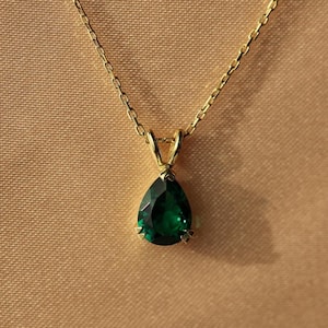 May include: A gold necklace featuring a pear-shaped, emerald-green gemstone pendant. The pendant hangs from a fine gold chain. The background is a plain, light-brown colour.