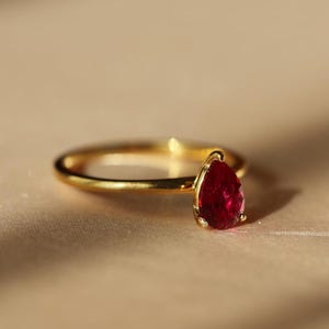 Pode incluir: Um anel de ouro com uma pedra preciosa vermelha escura em forma de pêra. O anel tem uma banda fina e polida, e a pedra preciosa é definida em uma delicada moldura de ouro. O anel é exibido em uma superfície de tom neutro, com iluminação suave.