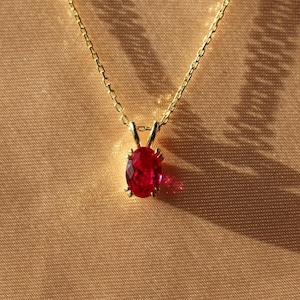 Könnte beinhalten: Eine goldfarbene Halskette mit einem kleinen, ovalen, roten Edelsteinanhänger. Die Kette ist filigran, und der Anhänger ist in einem goldfarbenen Rahmen gefasst. Der Hintergrund ist einfarbig hellbraun, mit einem Schatten der Kette.