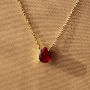 May include: A gold necklace with a pear-shaped, deep red gemstone pendant. The pendant hangs from a delicate gold chain. The necklace is set against a neutral, tan-coloured background.