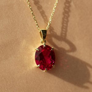 Könnte beinhalten: Eine goldfarbene Halskette mit einem großen, ovalförmigen, roten Edelsteinanhänger. Der Anhänger wird von vier Krappen gehalten. Die Kette ist filigran und goldfarben. Der Hintergrund ist einfarbig und neutral.