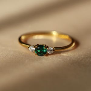 Può includere: Un anello d'oro con uno smeraldo rotondo centrale e due pietre rotonde e chiare più piccole. L'anello è posto su una superficie neutra, con un'illuminazione soffusa che ne evidenzia i dettagli.