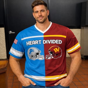 Puede incluir: Una camiseta azul y burdeos con cuello en V con el texto "HEART DIVIDED". La camiseta presenta un casco de fútbol a cada lado, uno plateado y otro burdeos. La manga izquierda tiene una bandera americana.