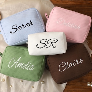 Pode incluir: Cinco nécessaires de cosméticos personalizados em cores pastel: azul claro, rosa, branco, verde e castanho. Cada nécessaire apresenta um nome bordado em uma cor contrastante. Os nécessaires são retangulares com fecho de zíper e uma pequena alça.