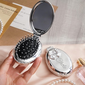May include: A silver compact mirror and hairbrush set, open to reveal a mirror and a black and white bristle brush. The closed compact has a bow design and the name "Chole" engraved on it. A pearl necklace and perfume bottle are also in the image.