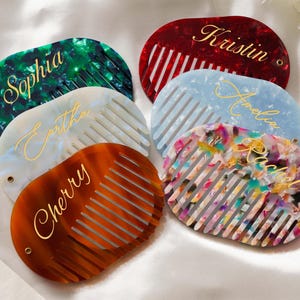 May include: A selection of personalised combs in shades of green, red, blue, and orange. Each comb displays a name in gold script. The combs are oval-shaped with evenly spaced teeth, and are made from a marbled material.