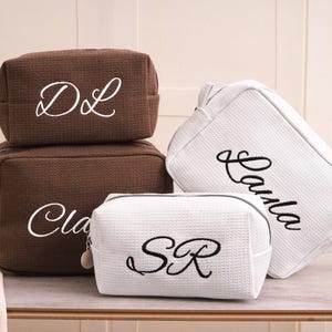 Può includere: Quattro trousse per cosmetici di diverse dimensioni e colori. Due borse marroni sono impilate, con iniziali ricamate bianche. Una borsa bianca con scritta ricamata nera è accanto a una borsa bianca più piccola con iniziali ricamate nere. Tutte hanno cerniere.