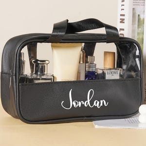 May include: Black cosmetic bag with a clear top, containing various beauty products. The bag has a handle and a front pocket with the name "Jordan" in white script. The bag is sitting on a light-colored surface.