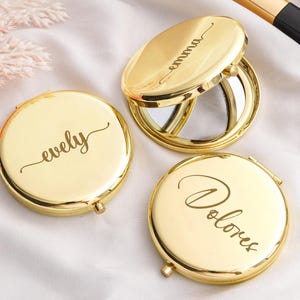 May include: Three gold-colored compact mirrors with personalized names engraved on them. One mirror is open, revealing a reflective surface. The names "evely", "emma", and "Dolores" are elegantly scripted on the compacts. Makeup brush and pearls are in the background.