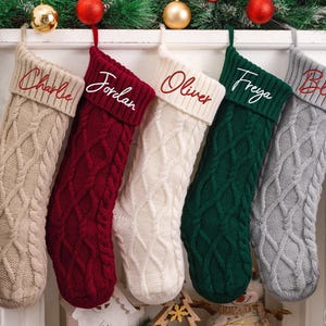 May include: Five cable-knit Christmas stockings in beige, red, white, green, and gray, each with a name embroidered in a contrasting color. Festive holiday decor with ornaments and greenery.