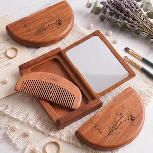 May include: A wooden compact mirror case with a hinged lid, featuring a laser-engraved poppy flower and the name Karen. Inside the case is a rectangular mirror and a matching wooden comb engraved with the name Karen. The wood has a warm brown tone.