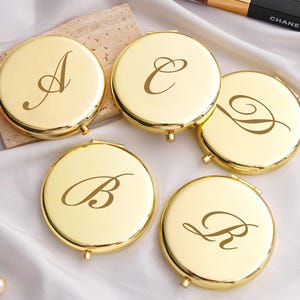 Può includere: Cinque specchietti compatti dorati, ciascuno con una lettera diversa in corsivo: A, B, C, D e R. Gli specchietti sono rotondi e hanno una finitura lucida. Un pennello per il trucco e un tubetto di mascara Chanel sono sullo sfondo.