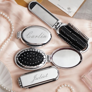 May include: Silver compact mirrors and a folding brush are displayed on a pink satin surface. Two mirrors are oval, one rectangular, and all have engraved names. A pearl necklace and perfume bottle are also in the image. The brush has black bristles.