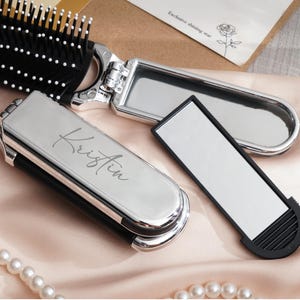 Peut inclure: Un miroir de poche argenté avec le nom "Kristin" gravé, une brosse à cheveux noire et argentée et un petit miroir rectangulaire. Les articles sont disposés sur une surface en satin rose avec un collier de perles.