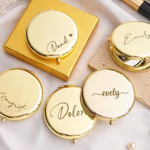 May include: Gold compact mirrors with personalized names in elegant script. The mirrors are round and open to reveal a mirror. Some are on a gold box and a stone block. A makeup brush and mascara are also visible.