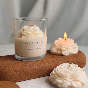 May include: A clear glass candle holder with a beige candle featuring a flower design. The candle is labeled "GALANNA CANDLES" and "HANDMADE | SOY WAX". Two additional flower-shaped candles are also visible, one lit with a flame. The candles are on a brown cork surface.