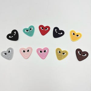 Peut inclure: Plusieurs écussons en forme de cœur de différentes couleurs, dont noir, rouge, turquoise, rose et jaune. Chaque écusson présente un motif d'œil stylisé. Les écussons sont disposés sur un fond blanc.