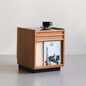 May include: A wooden side table with a black base and a magazine rack. A black coffee cup and saucer sit on top. The magazine in the rack has the letters "AD" on the cover. The table has a light brown finish.