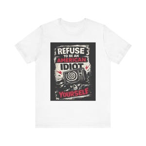 May include: White t-shirt with a graphic design. The design features the text "REFUSE TO BE AN AMERICAN IDIOT YOURSELF" in a distressed font, along with an image of a television with a skull and lightning bolts.