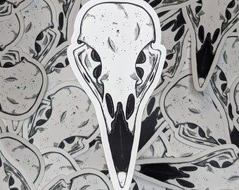 May include: A white sticker featuring a detailed black and white illustration of a bird skull. The skull is the focal point, with a prominent beak and eye sockets. The sticker is surrounded by other stickers with similar designs.