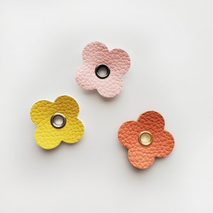 Puede incluir: Tres formas de flores de fieltro con un ojal de metal en el centro. Las flores son rosa, amarilla y naranja.