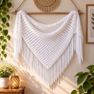 May include: A white triangular crochet shawl featuring a textured popcorn stitch pattern and long fringe along the bottom edges. The shawl is draped over a horizontal wooden rod, displaying its intricate knit design and symmetrical shape.