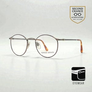 May include: Round, brown metal eyeglasses with clear lenses. The frames have a thin, delicate design with a subtle sheen. The temples feature a tortoiseshell pattern. The brand name "GIORGIO ARMANI" is visible on the lens. The image also includes the text "SECOND CHANCE EYEWEAR".