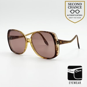 Puede incluir: Gafas de sol de gran tamaño con montura de carey marrón y lentes tintadas. La montura presenta una forma cuadrada redondeada con patillas finas. Una insignia en la esquina superior derecha dice SECOND CHANCE AUTHENTICATORS VERIFY THE VISION.