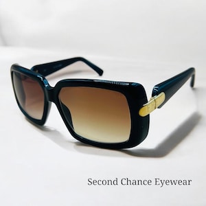 Op de afbeelding: Donker marineblauwe zonnebril met een rechthoekig montuur en bruine verloopglazen. De veren hebben een gouden accent. De tekst "Second Chance Eyewear" staat onderaan de afbeelding.