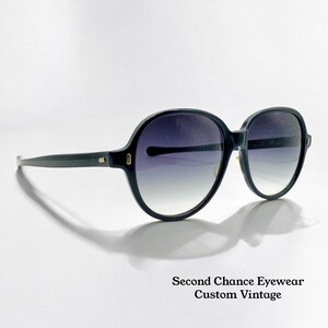 May include: Black oversized sunglasses with gradient lenses. The round frames have a dark finish, with a subtle gold accent near the hinges. The text "Second Chance Eyewear Custom Vintage" is at the bottom.