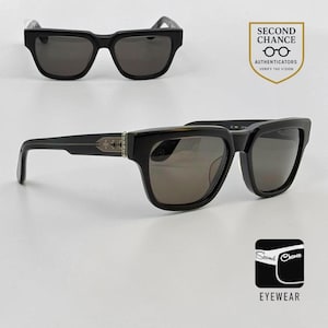 May include: Black sunglasses with dark tinted lenses and thick frames. The temples feature a metallic silver-toned inlay with a cross motif. The image includes a logo with the text SECOND CHANCE AUTHENTICATORS and a logo with the text SECOND CHANCE EYEWEAR.
