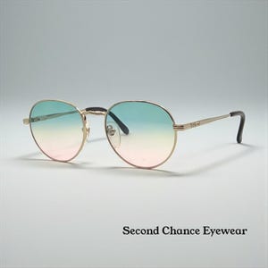 Puede incluir: Gafas de sol redondas con montura dorada y lentes degradadas, que van del rosa al azul. El diseño es clásico, con varillas finas y metálicas. El texto "Second Chance Eyewear" es visible en la parte inferior.